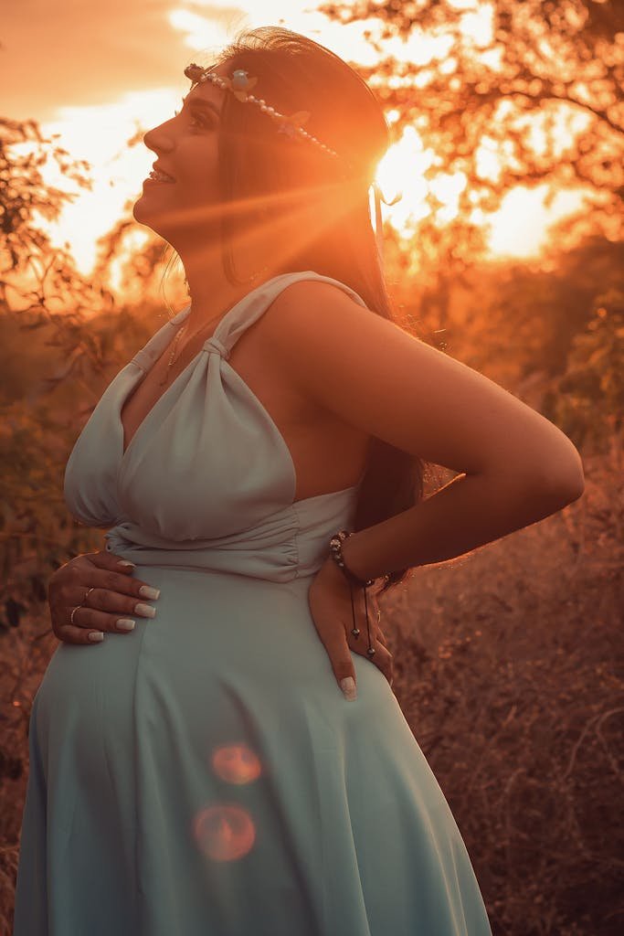 Side view of happy pregnant female standing with hand on back and on belly in nature with sun shining through branches of tree